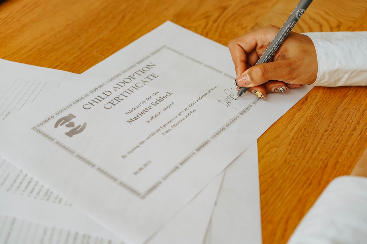 A close-up of a hand signing a child adoption certificate with a pen on a wooden table.